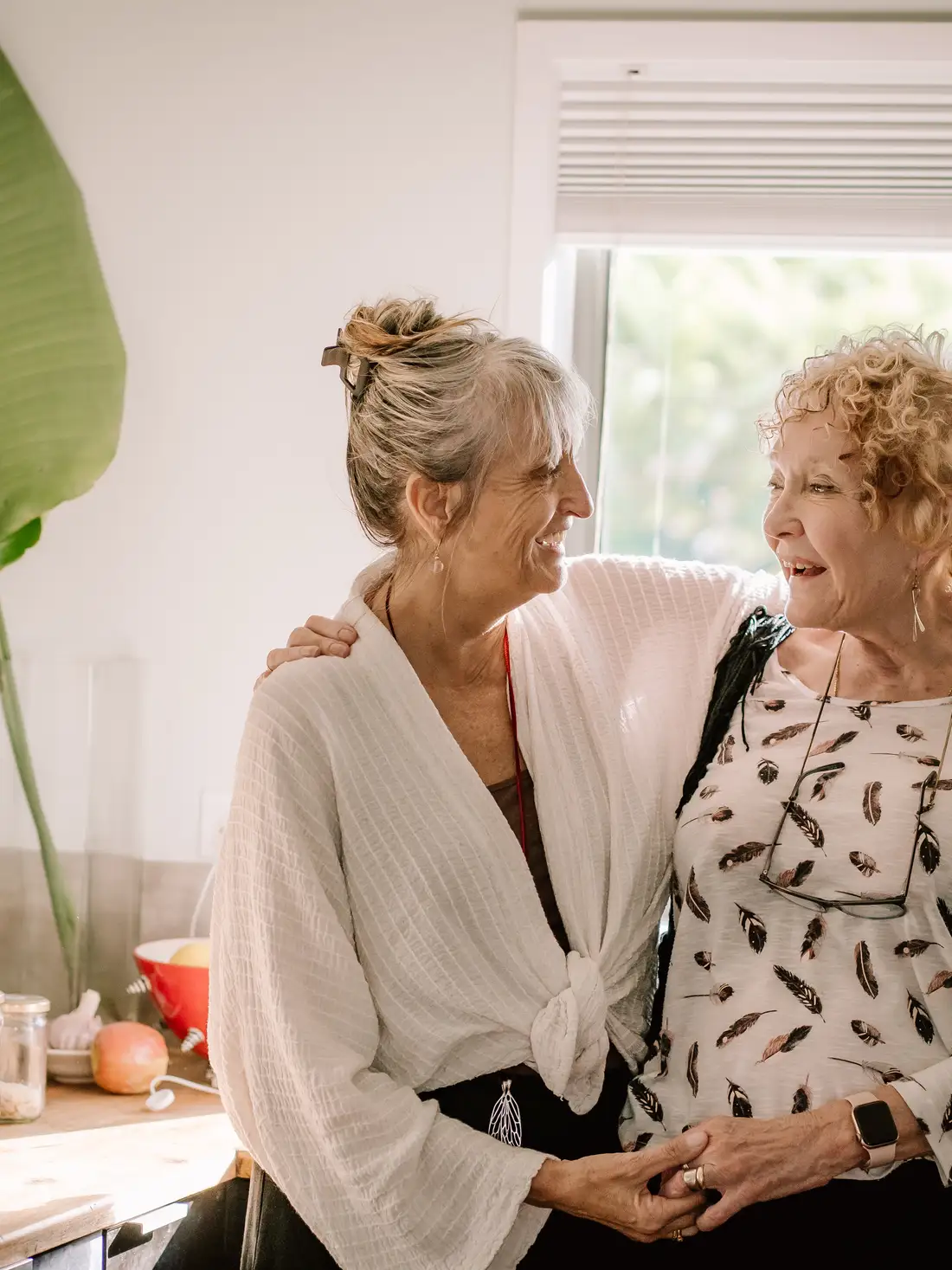 Two women smiling and embracing in a sunlit kitchen