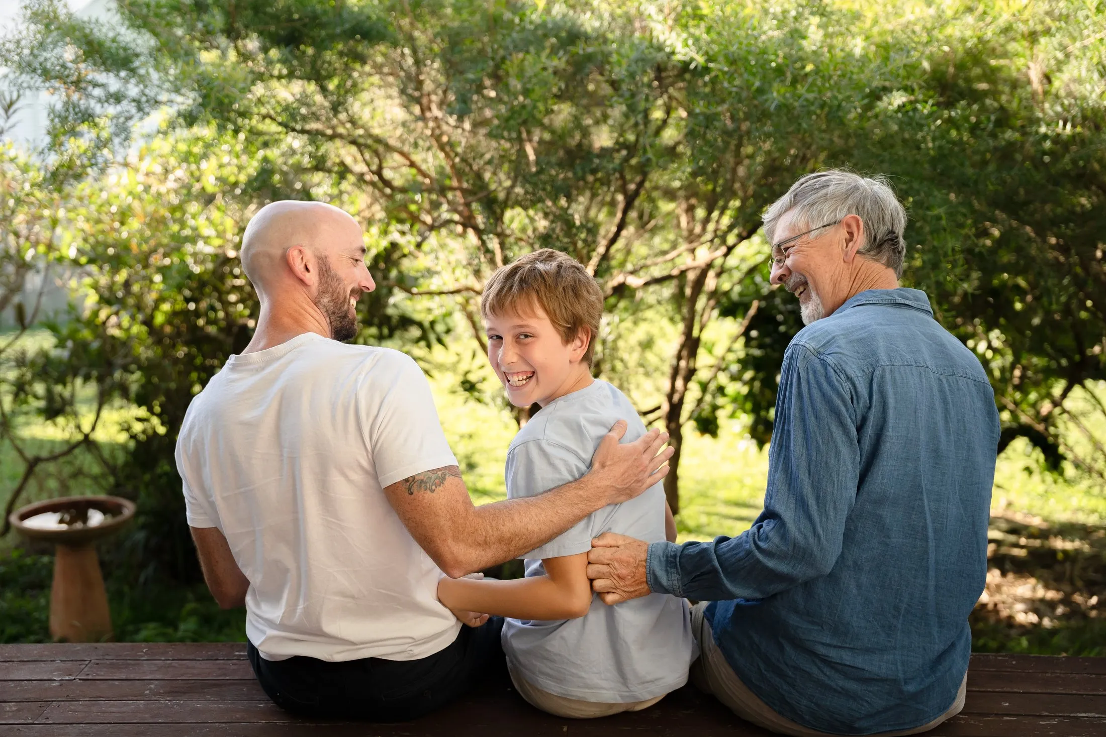 A grandmother, grandchild and grandfather sitting close together on a garden bench in warm golden afternoon light