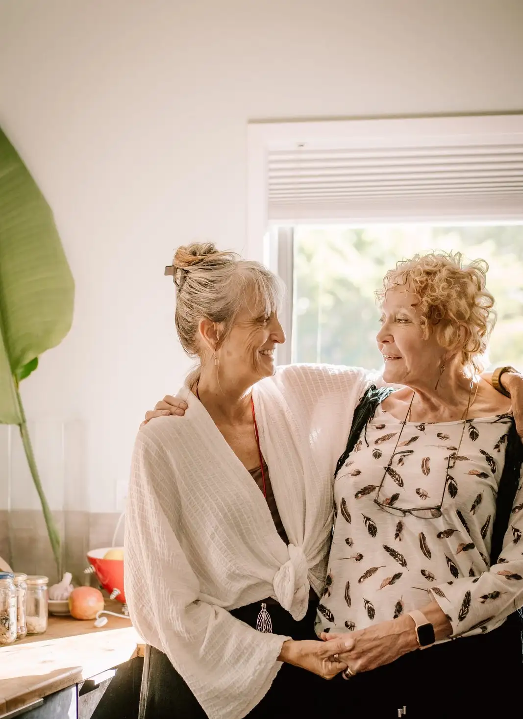 Two women smiling warmly with arms around each other in a sunlit kitchen